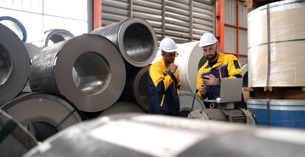 Medewerkers in een fabriekshal bekijken productiegegevens tussen grote opgerolde metalen platen.
