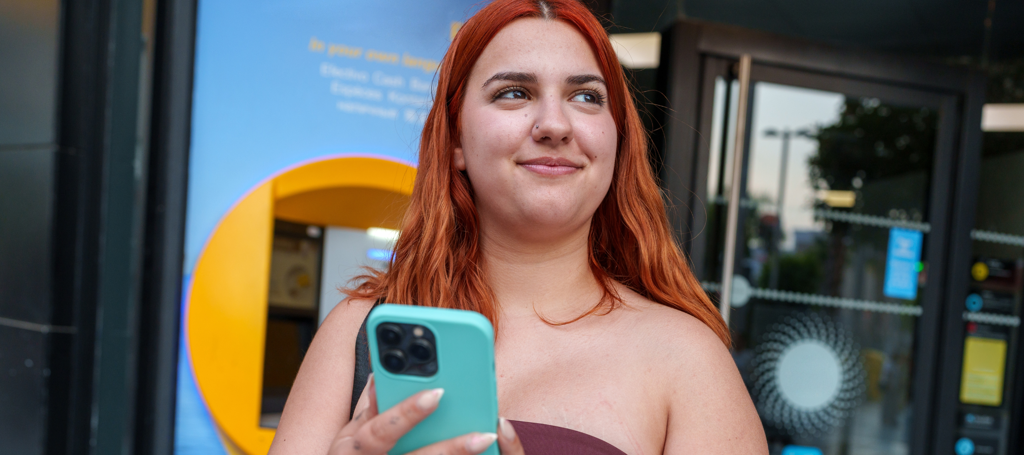Meisje staat met haar telefoon in haar hand voor een pinautomaat