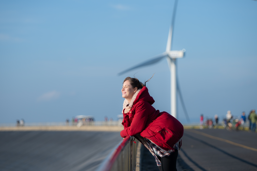 Glimlachende vrouw met ogen dicht in de wind met op de achtergrond een windmolen.