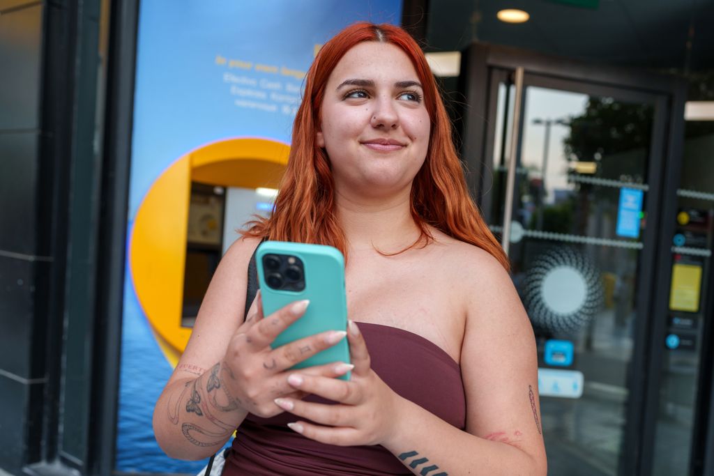 Meisje staat met mobiele telefoon in haar hand voor een pinautomaat